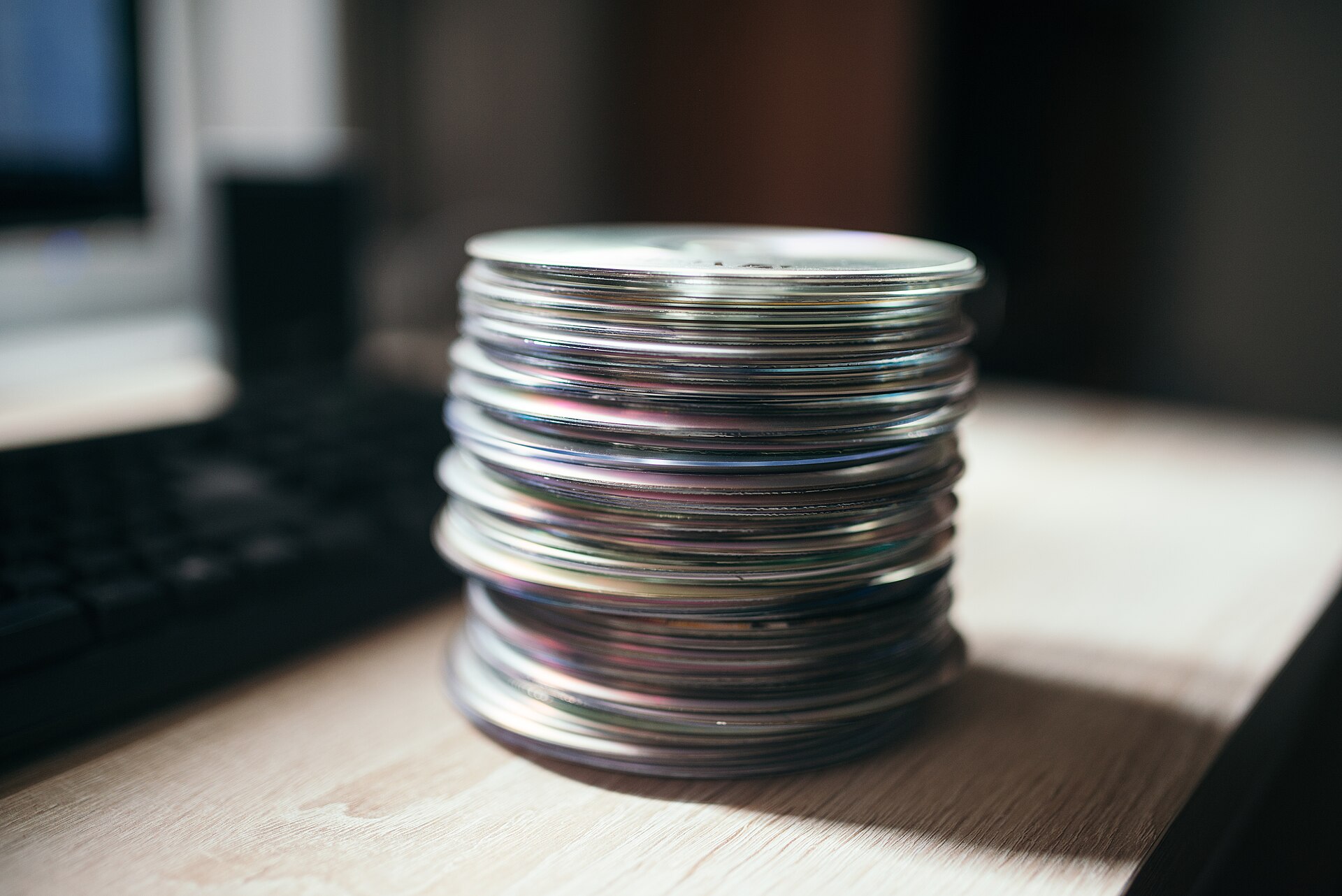 A stack of compact discs resting on a wooden table beside a computer keyboard.