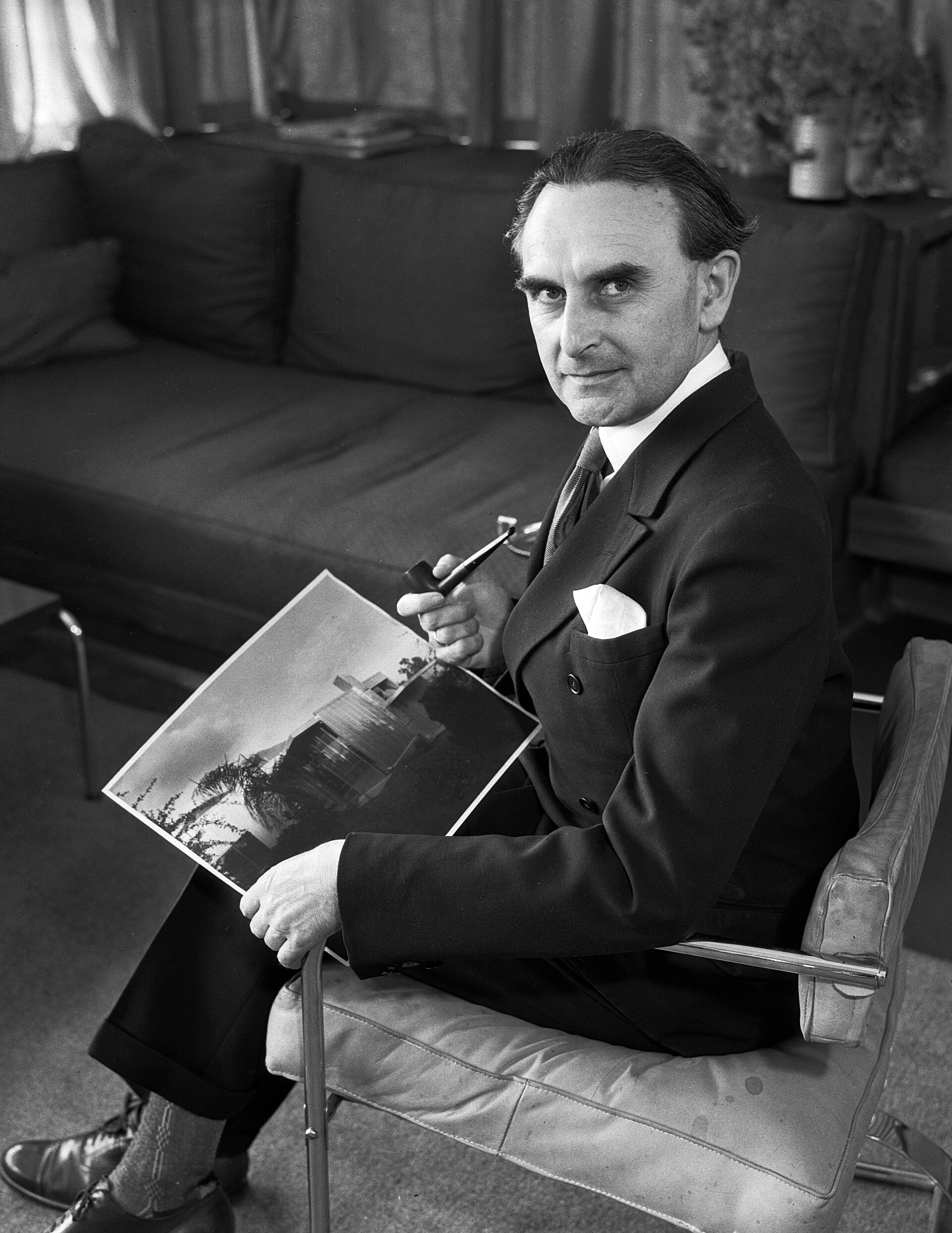 Richard J. Neutra, seated in a living area on a cantilever chair he designed, holding a pipe and a photograph of the William and Melba Beard House in Altadena, California, for which he won a gold meda